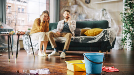 Panicing Couple In Despair Sitting on a Sofa Watching How Water Drips into Buckets in their Living Room