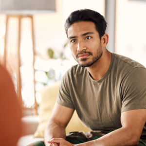 Man sitting on a couch looking concerned. Another persons shoulder is out of foucs in front of the camera, they are facing the man.