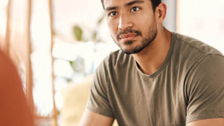 Man sitting on a couch looking concerned. Another persons shoulder is out of foucs in front of the camera, they are facing the man.