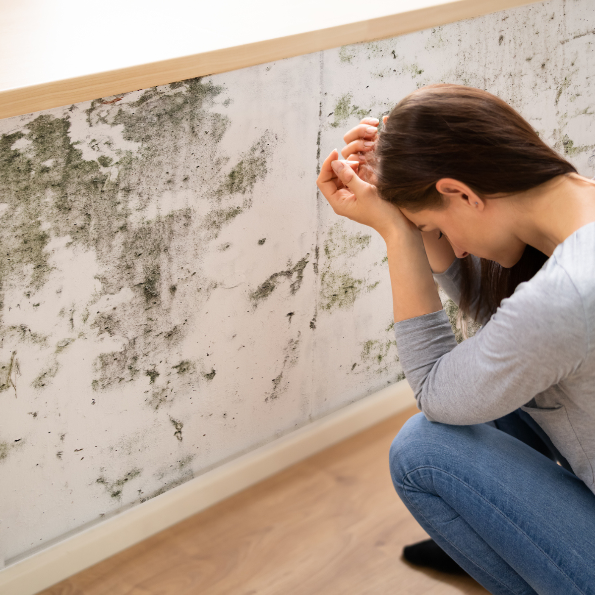 Woman crouching beside an indoor wall covered in dark mold patches, with hands resting on forehead.