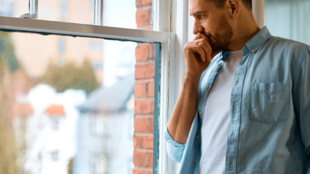 Person standing indoors by a large window, looking outside with a hand resting against the chin. The person is wearing a light blue shirt over a white T-shirt and neutral-coloured trousers. Natural light comes through the window, showing a blurred view of buildings outside and a brick wall along the window frame.
