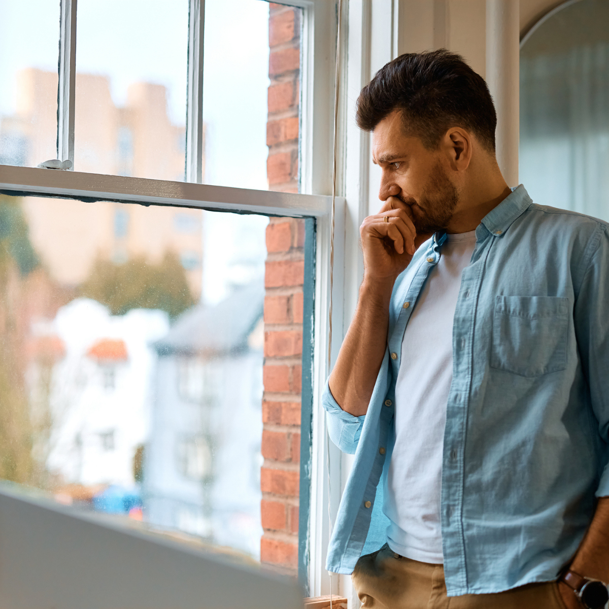 Person standing indoors by a large window, looking outside with a hand resting against the chin. The person is wearing a light blue shirt over a white T-shirt and neutral-coloured trousers. Natural light comes through the window, showing a blurred view of buildings outside and a brick wall along the window frame.