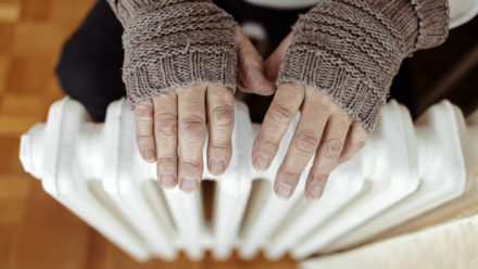 A man hands in wool gloves warm near the heater. Old men's hands in knitted gloves on heating radiator at home during the day. Person heating their hands at home over a domestic radiator in winter.